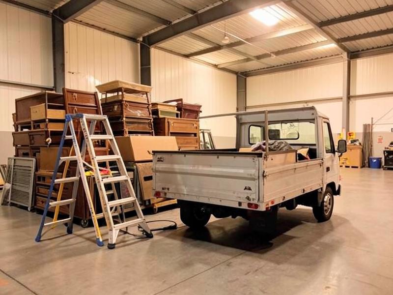 Ladders, tools and a utility truck beside stacked furniture in a warehouse